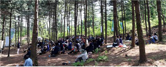 사람들이 숲속에 편하게 앉아서 강의를 듣고있습니다.